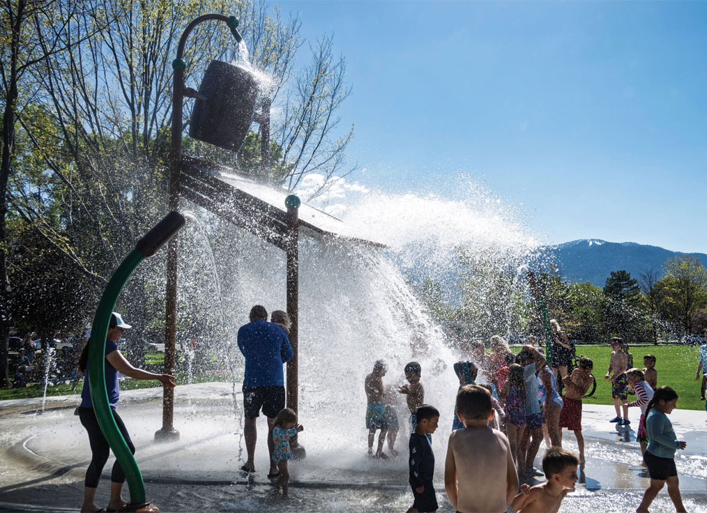 a crow of people gathers under a massive splash of water from a large dumping bucket water feature on a splash pad