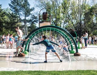 child interacting with jets on the entrance of a splash pad that features three green and brown water rings and a large dumping bucket