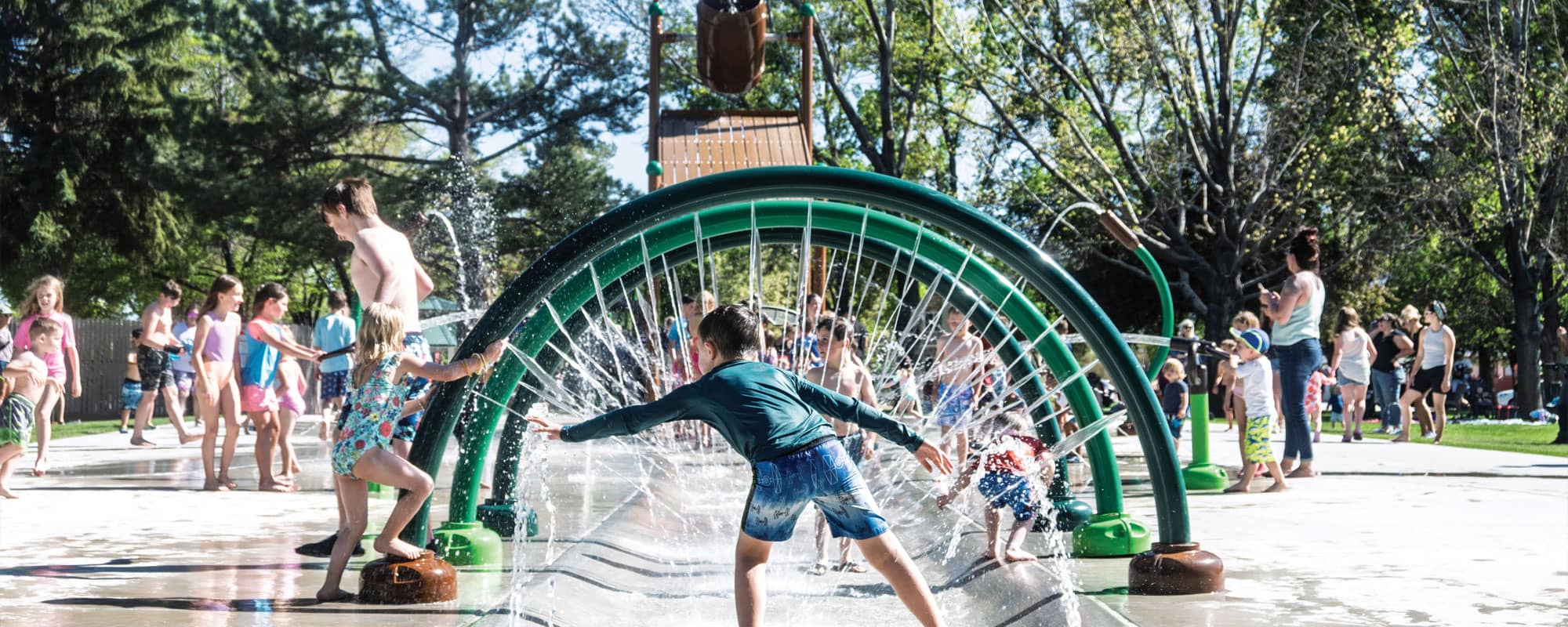 child interacting with jets on the entrance of a splash pad that features three green and brown water rings and a large dumping bucket