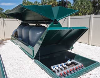 a green sand filtration cabinet with grey sand filters and a green valve box in an outdoor equipment yard that is gated