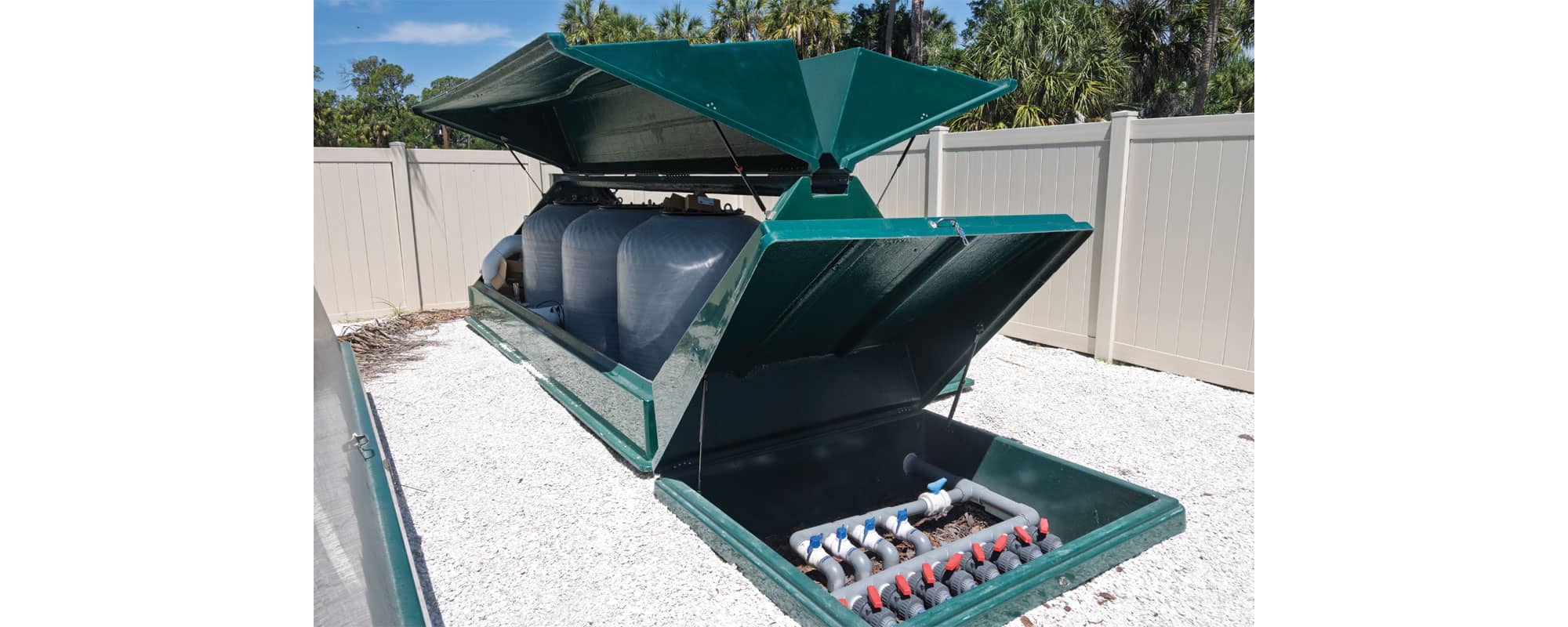a green sand filtration cabinet with grey sand filters and a green valve box in an outdoor equipment yard that is gated