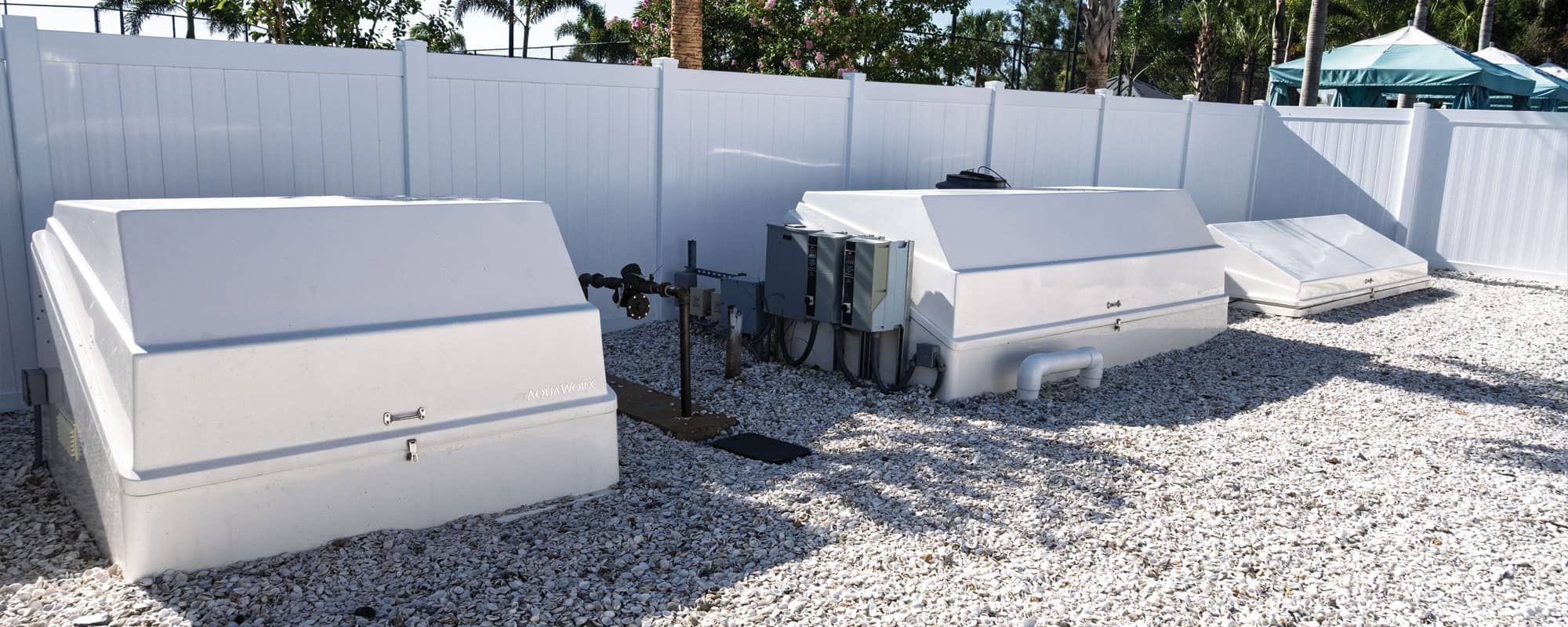 white swimming pool filtration equipment packs are housed in a gated, outdoor equipment yard